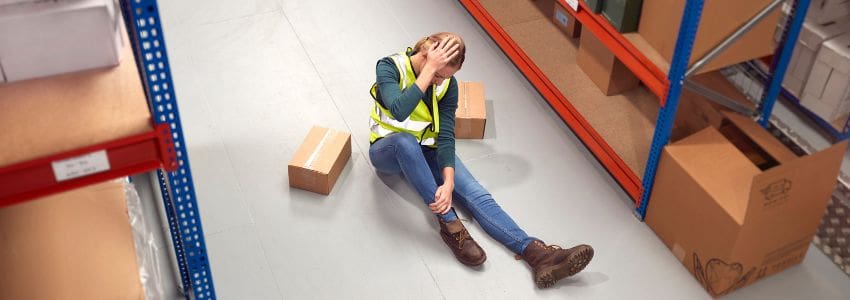 Female warehouse worker sitting on the ground holding her head and ankle with boxes laying askew next to her.