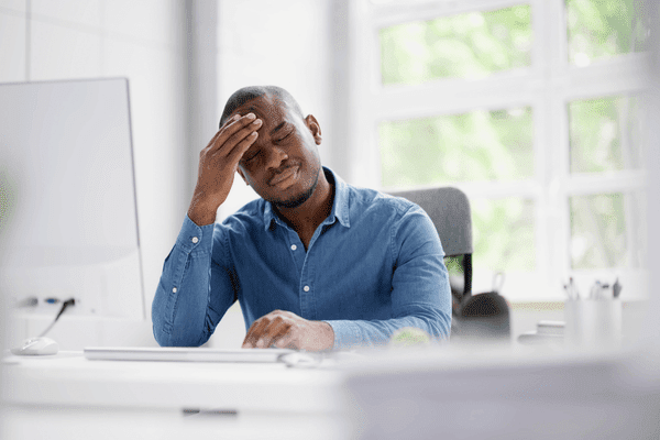 Stressed businessman sitting at his desk in front of his computer with his eyes closed and his hand rubbing his forehead.